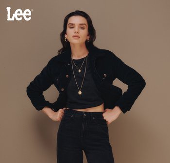 Vrouw in een zwarte cropped jack en hoge taille zwarte jeans, met gouden creolen en gelaagde gouden kettingen, staande tegen een beige achtergrond.