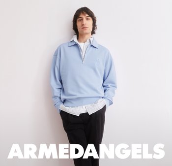 Giovane uomo con felpa azzurro chiaro con cerniera a quattro quarti sopra una camicia a quadri, in piedi rilassato contro un muro bianco con il testo "ARMEDANGELS" sotto.