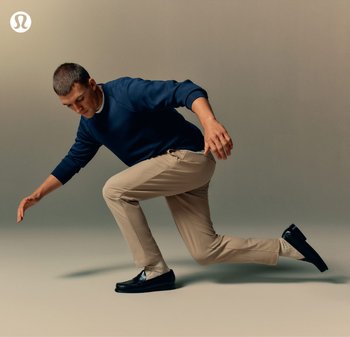 Uomo con maglione blu navy, pantaloni beige e mocassini neri che si equilibra basso con un ginocchio piegato e una gamba distesa su uno sfondo beige semplice.