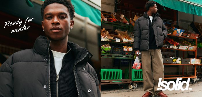 Schwarze Pufferjacke mit hohem Kragen, kombiniert mit einem dunklen Pullover und hellbeigen Hosen. Der Hintergrund zeigt einen Obstmarkt mit grünen Akzenten.