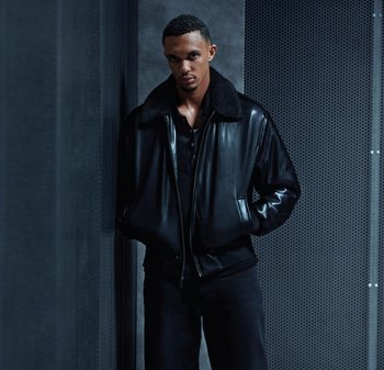 Svart läderbomberjacka med en fårskinnskrage, kombinerad med en svart skjorta med knappar. Modellen poserar mot en strukturerad bakgrund.