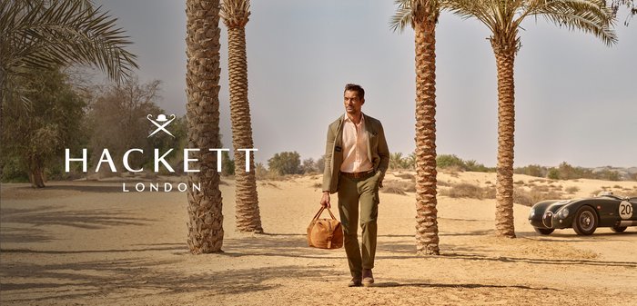 Hombre con traje verde que lleva un bolso de cuero beige caminando entre palmeras en un paisaje desértico con un coche antiguo cerca.