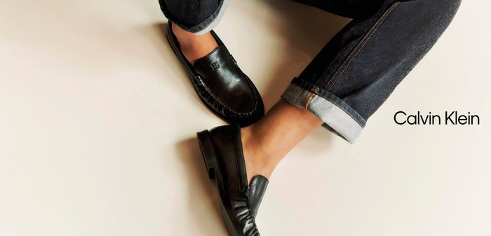 Black leather loafers worn with cuffed dark jeans on a light background, with Calvin Klein brand name visible.