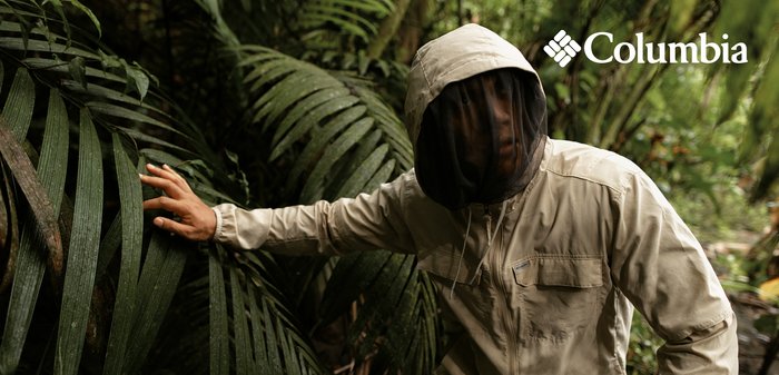 Person wearing beige insect-repellent jacket with hood and black mosquito net, navigating dense green jungle foliage outdoors.
