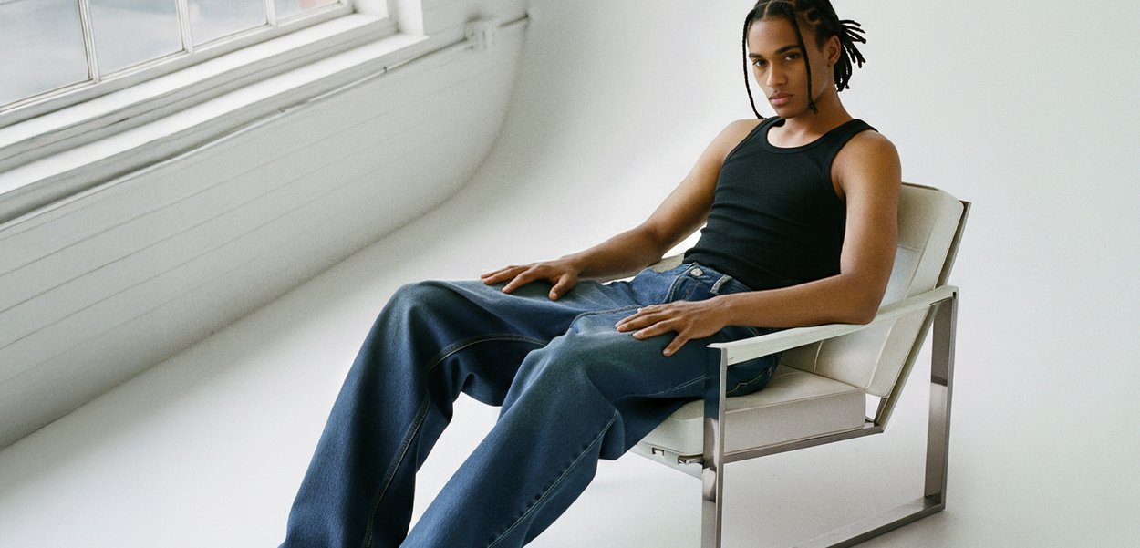 Svart tanktop, løse blå denimjeans, sitter i en hvit, moderne stol med metallramme. Naturlig lys fra et vindu i bakgrunnen.