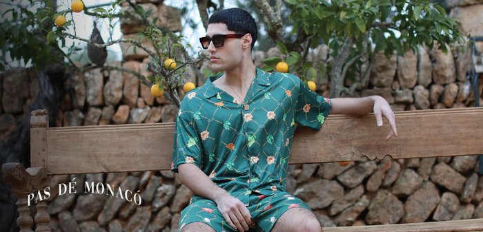 Grønn kortermet skjorte og matchende shorts med blomstermønster og frukt. Bært med solbriller, sittende på en trebänk.