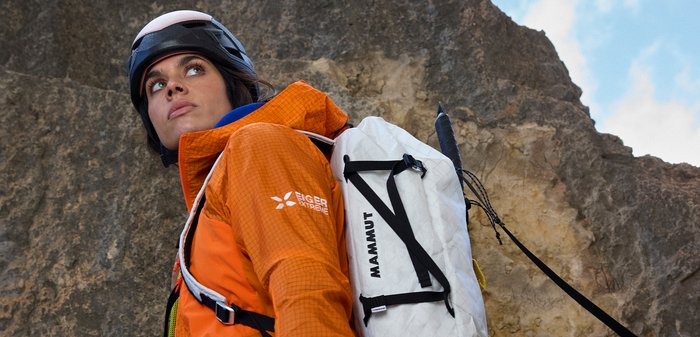 Beklimmer met oranje jas en helm, draagt een witte Mammut-rugzak, kijkt omhoog tegen een rotsachtig berglandschap.