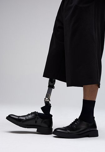 Scarpe formali in pelle nera con lacci, abbinate a pantaloncini neri fino al ginocchio. Una gamba protesica è visibile, con un giunto metallico e un calzino nero.