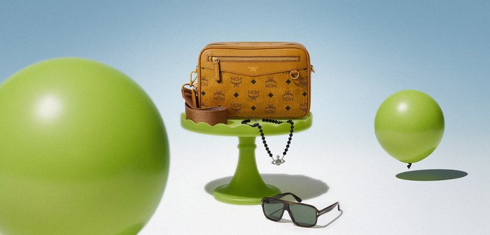 Rjava oblikovalska torbica čez ramo na zelenem podstavku, ob njej črna sončna očala in ogrlica z biseri, v ozadju plavajo dva zelena balona.