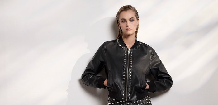 Giacca bomber in pelle nera con borchie argentate sul davanti, chiusura con zip, polsini e orlo a coste, texture liscia con una forma aderente.