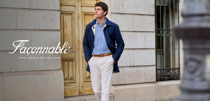 Jonge man die dicht bij een stenen gebouw wandelt, draagt een blauw jasje, gestreept hemd, beige broek en loafers, handen in zijn zakken, kijkt opzij.