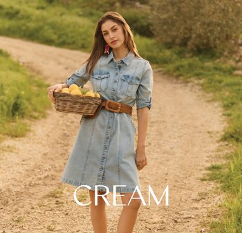 Woman standing on a dirt path holding a basket of lemons, wearing a light denim dress with rolled sleeves and a woven belt.