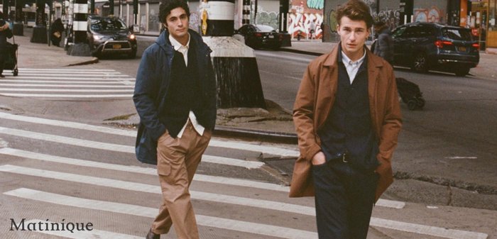 Two men walk on a city crosswalk. One wears a blue coat and tan pants, the other a brown jacket over a navy vest and dark pants.