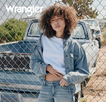 Ung person med krøllete hår iført denimjakke og jeans, stående foran et gjerde av lenketråd og en pickup lastebil utendørs.