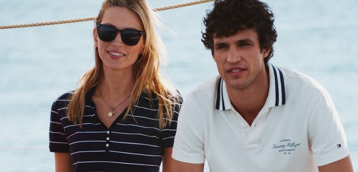 Deux jeunes adultes, une femme en lunettes de soleil et chemise rayée, et un homme en polo blanc, assis à l'extérieur près de l'eau.