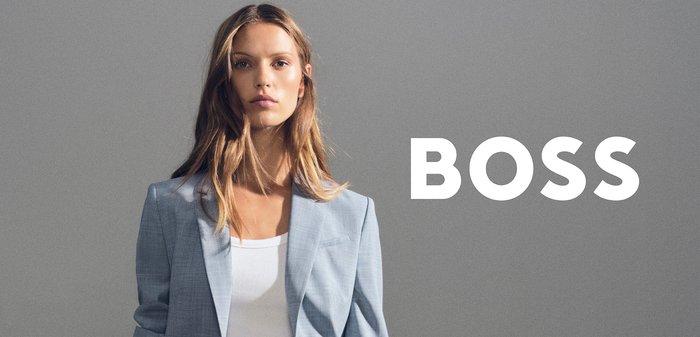 Young woman confidently posing in light blue suit and white top against gray background with large white "BOSS" text overlay.