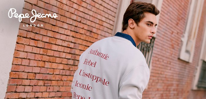 Sweatshirt branca com texto vermelho que diz "Authentic", "Rebel", "Unstoppable" e "Iconic". Apresenta uma gola de ganga azul num cenário com parede de tijolos.