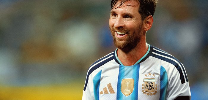 Joueur de football masculin souriant avec barbe, portant le maillot rayé bleu et blanc de l'Argentine avec les logos de la FIFA et de l'AFA sur le terrain.