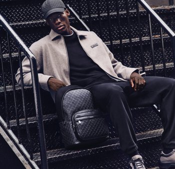 Ung man i grå jacka och svart keps sitter på metalltrappa med en texturerad svart ryggsäck, iförd svarta byxor och sneakers.