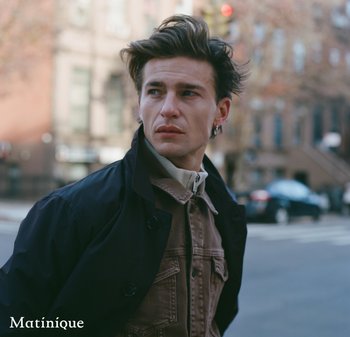 Young man with tousled hair and earring wears layered jackets, looking sideways on a city street with blurred buildings and cars behind.