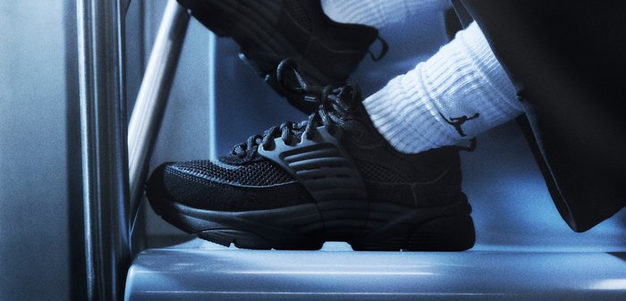 Personne portant des chaussures de sport noires et des chaussettes blanches côtelées avec un logo noir, assise avec les pieds posés sur une surface métallique.