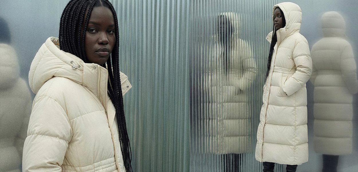 Roomkleurige donzen jas met hoge kraag en kap, met horizontale stiksels en zijzakken, gedragen over zwarte broek.