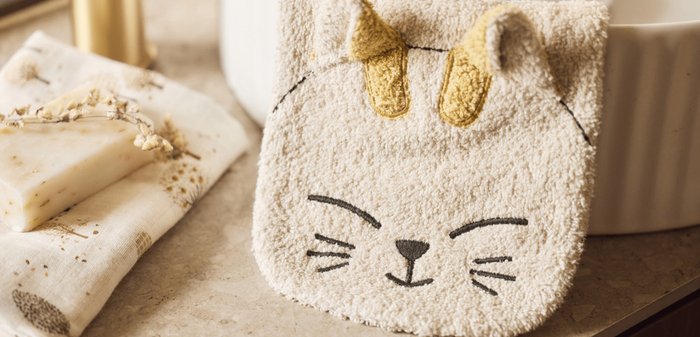 Handdoek in de vorm van een kattengezicht, gemaakt van zacht beige stof met gestikte kenmerken, waaronder zwarte snorharen en gele oren.