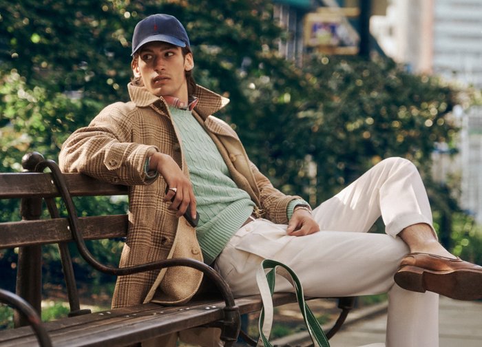 Joven con abrigo beige, suéter verde, pantalones blancos y gorra descansando en un banco del parque en un entorno urbano al aire libre.