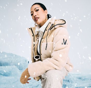 Beige und creme gepolsterte Jacke mit einem Kunstfell-Textur, schwarzen Reißverschlüssen und einem 'M'-Logo am Ärmel. Getragen mit einem weißen gerippten Kragen.