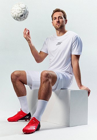 Man in witte sportkledij en rode voetbalschoenen zit op een wit blok en balanceert met één hand een voetbal in de lucht.