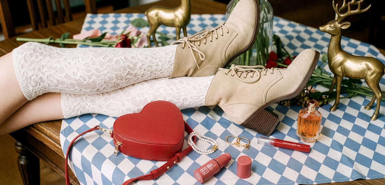 Jambes en chaussettes blanches en dentelle et bottines beige à talons reposent sur une nappe bleue et blanche avec un sac rouge en forme de cœur, des bijoux, un parfum et des figurines de cerfs en laiton.