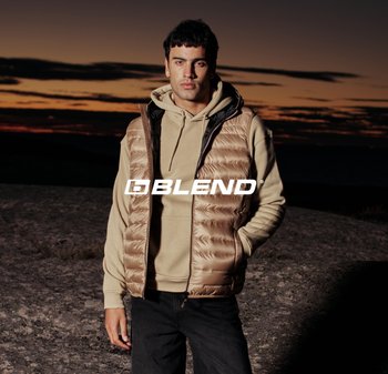 Mann trägt beige Kapuzenpullover und hellbraune gesteppte Weste, steht bei Sonnenuntergang auf felsigem Gelände vor dunklem Himmel, Logo "BLEND" in der Mitte.