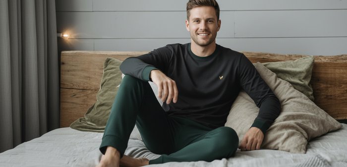 Smiling man wearing black long-sleeve shirt and green pants, sitting barefoot on a bed with pillows and wooden headboard behind him.