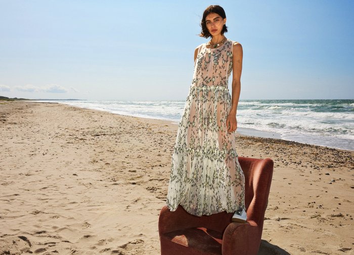 Mujer con un vestido floral translúcido y zapatillas, de pie sobre un sillón rojo en una playa arenosa con olas y cielo despejado de fondo.