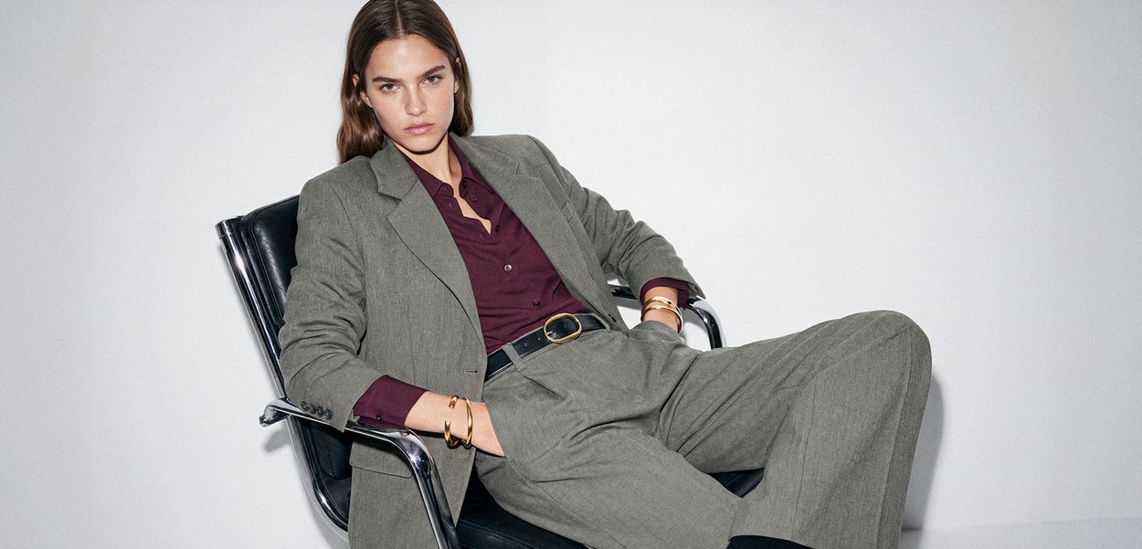 Pikajuukseline noor naine lamab mustal toolil, kandes halli ülikonda ja burgundia särki, vaadates otse kaamerasse.