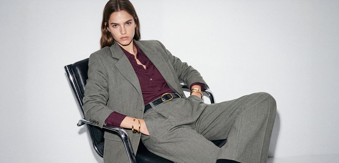 Νεαρή γυναίκα με μακριά μαλλιά που recline σε μαύρη καρέκλα, φορώντας γκρι κοστούμι και μπορντό πουκάμισο, κοιτάζει κατευθείαν την κάμερα.