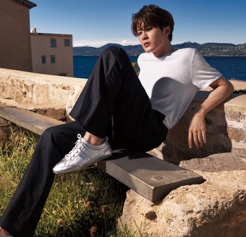 Junger Mann in weißem Hemd und schwarzer Hose sitzt auf einer Steinbank und blickt auf das Meer und ferne Hügel unter klarem Himmel.