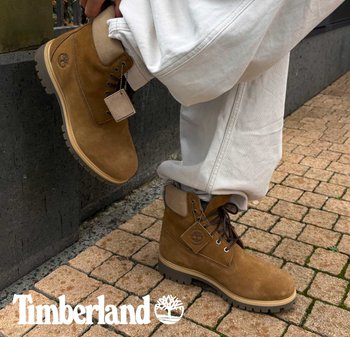 Braune Wildleder-Wanderschuhe mit Gummisohle, cremefarbenen Akzenten, runden Schnürsenkeln und einem Markenzeichen-Label, vor einem Kopfsteinpflaster-Hintergrund.