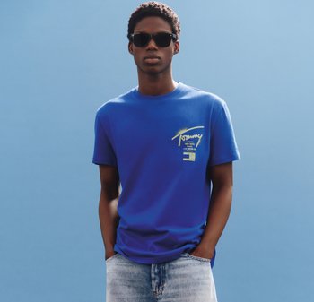 Jonge man met zwarte zonnebril, een blauw T-shirt met print en lichtblauwe jeans, staand tegen een effen blauwe achtergrond.