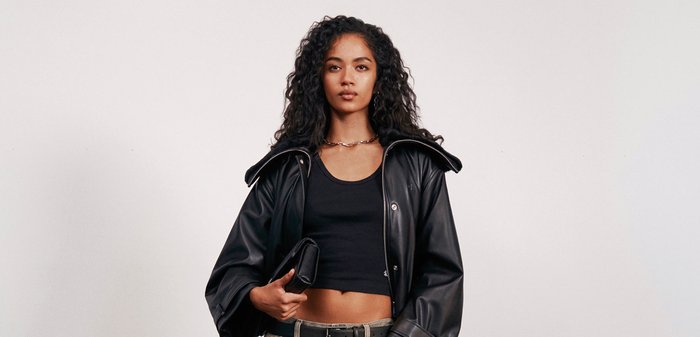 Young woman with curly hair wears a black cropped top, leather jacket, wide-leg pants, and sandals, standing in front of a black sofa.