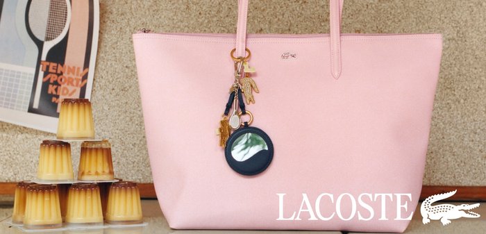 Bolso Lacoste rosa claro con accesorios de llavero dorados y negros sobre una superficie beige cerca de postres flan de caramelo apilados.