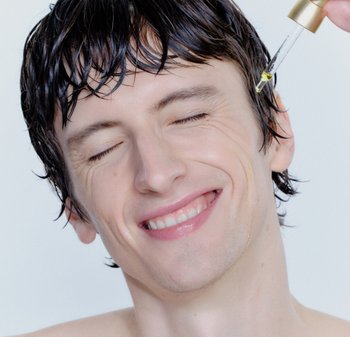 Compte-gouttes transparent déposant de l'huile jaune sur le visage d'une personne souriante, aux cheveux humides et à la peau lisse, sur un fond clair.