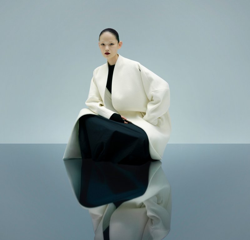 White oversized coat over a black dress, seated on a reflective surface. Smooth fabrics, minimalist design, and neutral colour palette.