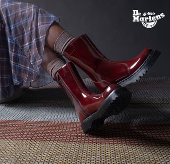 Jambes croisées portant des bottes Dr. Martens en cuir bordeaux brillant à semelles épaisses, des chaussettes marron et une jupe à carreaux bleus sur un tapis à motifs.