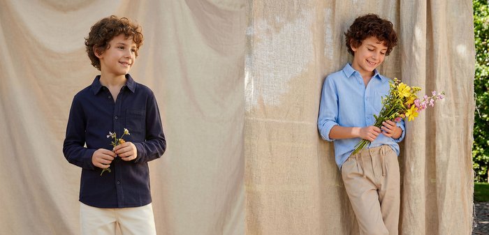 Enfant souriant portant une chemise bleue et un pantalon beige, tenant un bouquet de fleurs jaunes et roses, debout devant un rideau beige en extérieur.