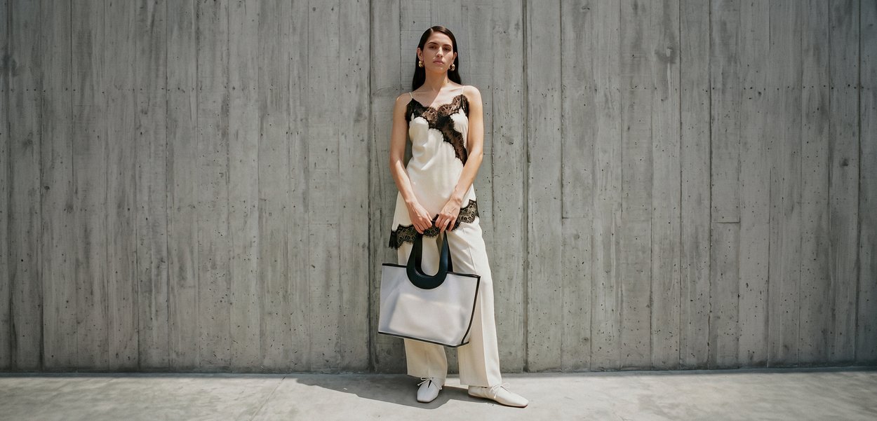 Mujer con conjunto blanco con detalles de encaje negro, de pie sosteniendo un gran bolso tote blanco y negro contra un fondo de pared de concreto gris.