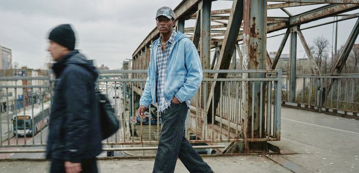 Jeune homme en veste en jean et casquette marchant sur un pont métallique rouillé avec un piéton flou qui passe dans un environnement urbain.