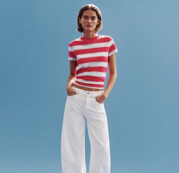 Vrouw staand met handen in de zakken, gekleed in een rood-wit-blauw gestreept t-shirt en wijde witte broek, voor een blauwe achtergrond.