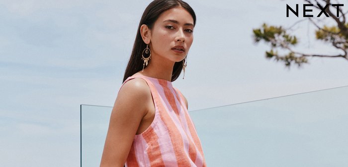 Pikkade tumedate juustega naine, kes kannab varrukateta roosa-oranži vertikaaltriibulist topi ja sobivat seelikut, seisab õues klaaspiirde kõrval.