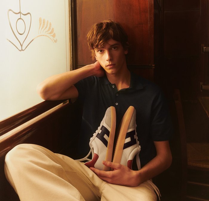 Giovane uomo con polo scura e pantaloni beige seduto vicino a una finestra, che tiene un paio di scarpe da ginnastica bianche e blu navy con suole in gomma.